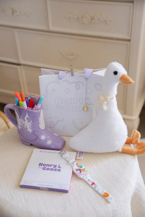 A plush Henry the Goose toy sits beside a branded gift bag, non-toxic fabric markers, and a creatively shaped container filled with colorful markers.